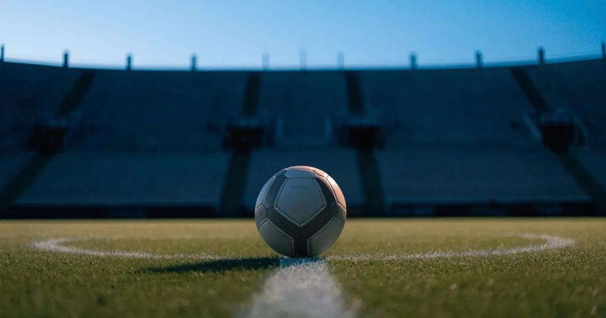 Ein Fussball liegt auf dem Mittelpunkt eines leeren Rasens, im Hintergrund steigen steile Tribünen in der Dämmerung auf.