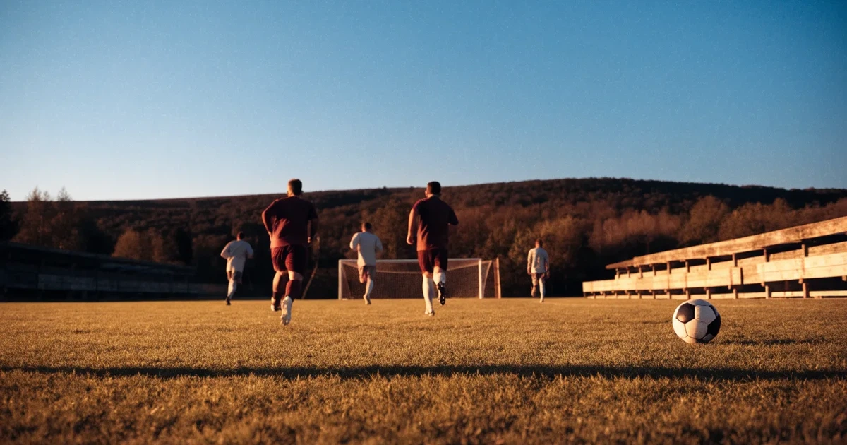 Ein kleiner Fussballplatz mit einfacher Holztribüne und Rasenstreifen am Rand einer Ortschaft, im Hintergrund ein bewaldeter Hügel im Abendlicht.