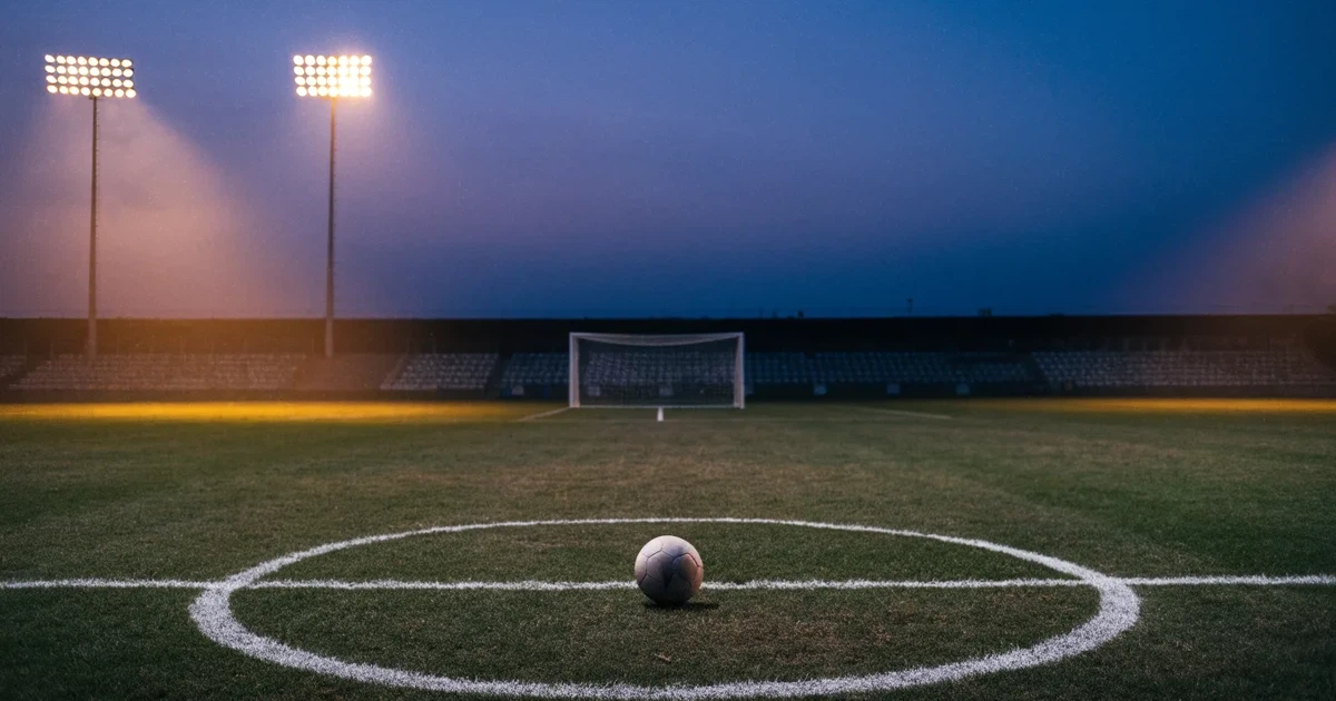 Ein Fussball liegt auf dem Anstosspunkt eines leeren Rasenplatzes in der Abenddämmerung, dahinter leuchten Flutlichtmasten über einer Tribüne.