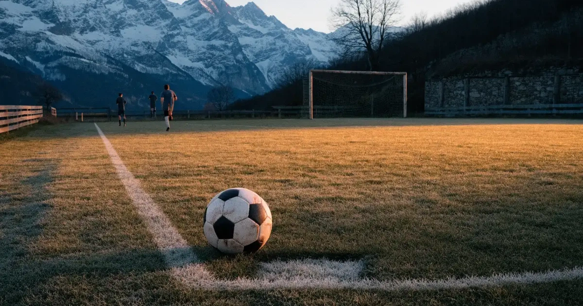 Ein Dorf-Sportplatz mit einem Ball am Strafraumrand, dahinter schneebedeckte Alpengipfel im klaren Morgenlicht