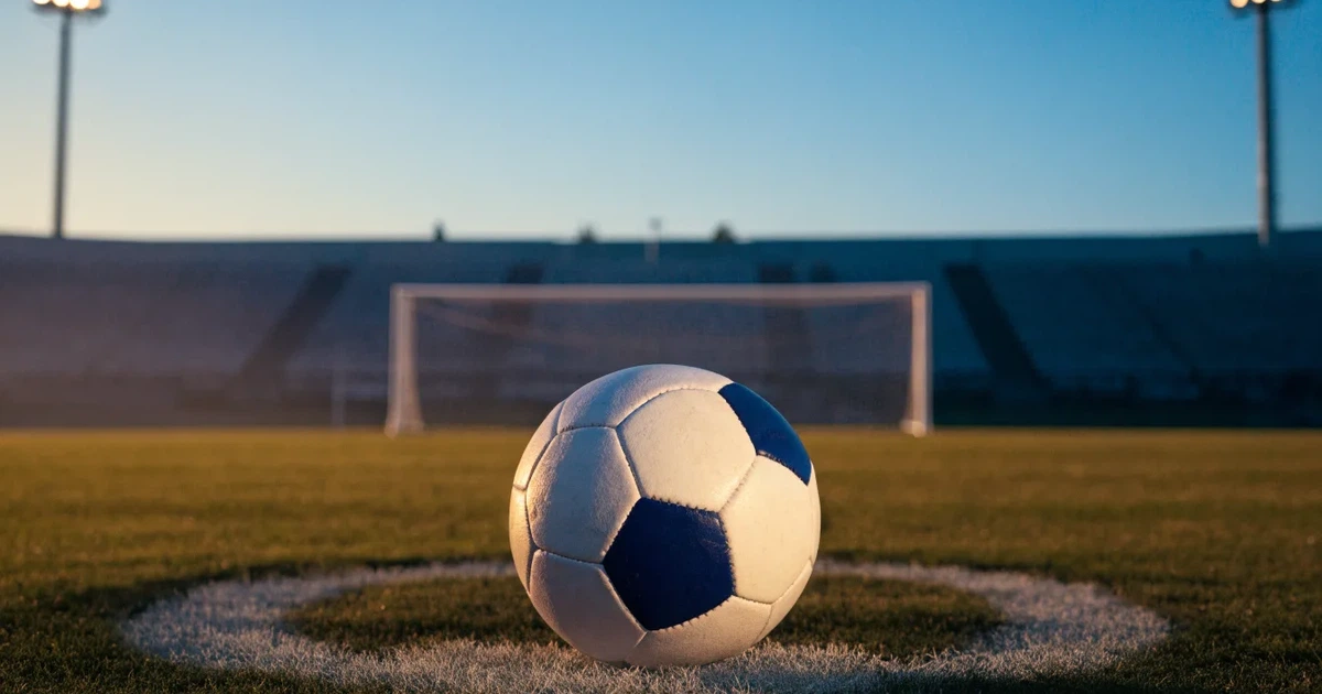 Ein Fussball liegt auf dem Anstosspunkt eines leeren Rasenplatzes, im Hintergrund glimmen Flutlichtmasten im Abendlicht.